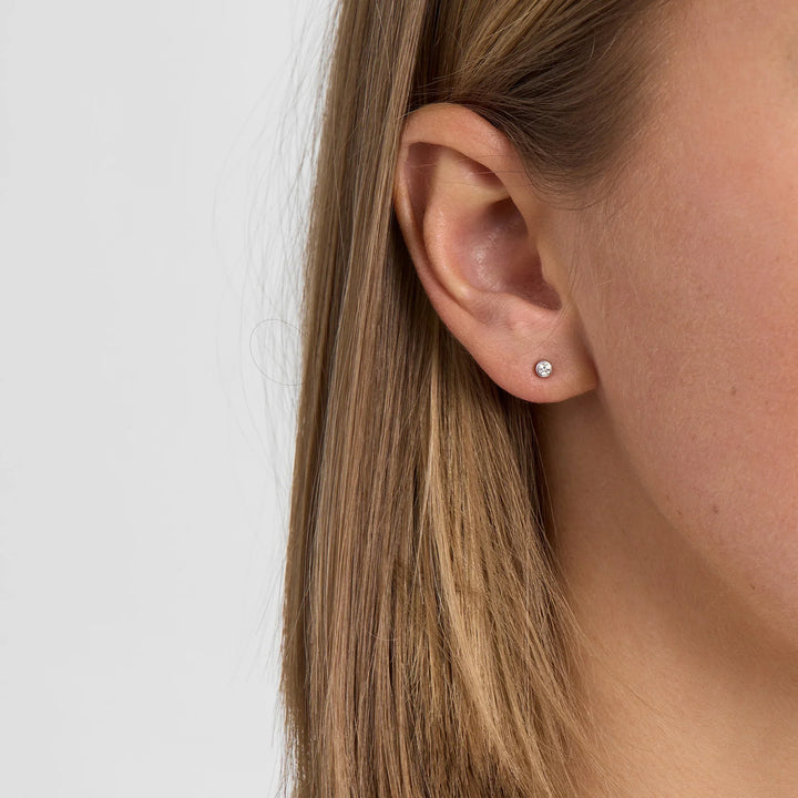 Earring in silver colored metal with single clear stone, shown on the ear from the side