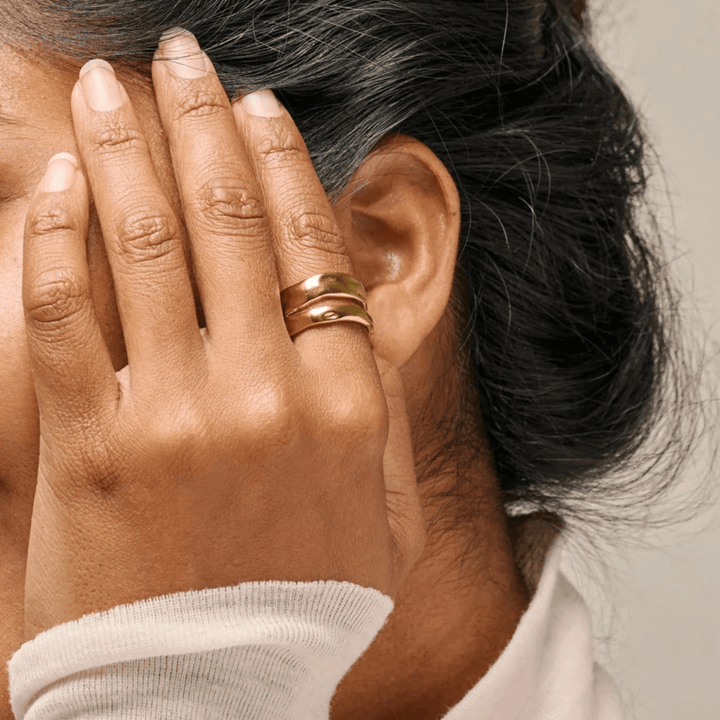 Enamel Sandra Statement Ring gold-plated appears on the woman's hand close to the ear, shiny golden surface. Model R92G-R92G-001.