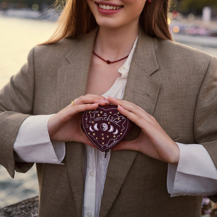 The Moonstone Heart Jewelry Box Maroon is shown in the hands of a woman, heart-shaped with embroidery, outdoors. Model 2026-4207-2