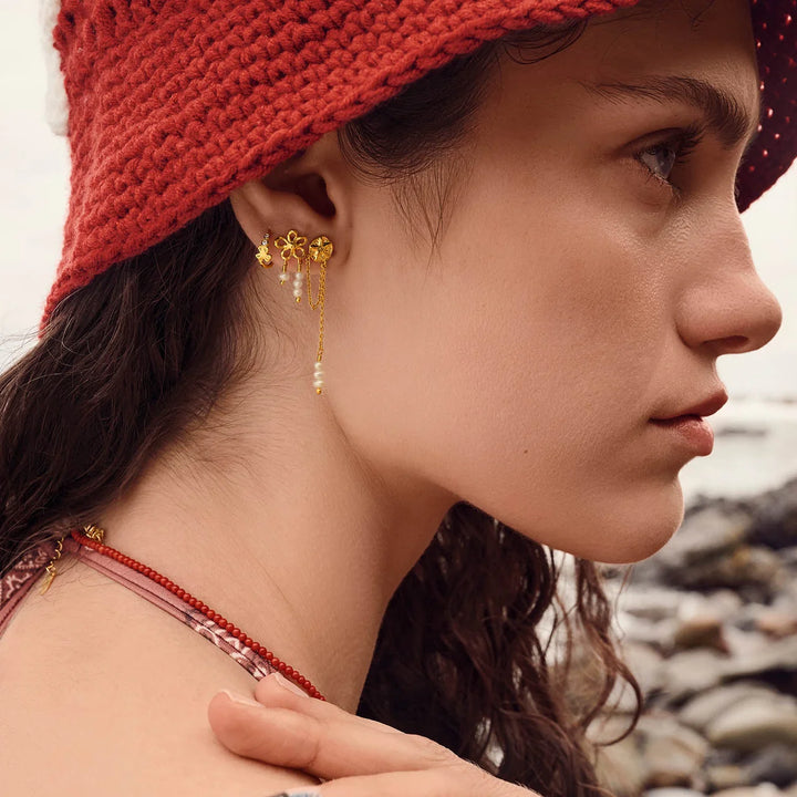 Maanesten Nicola gold-plated flower earrings with pearl details are seen on a model at the beach. Model 10106a-2