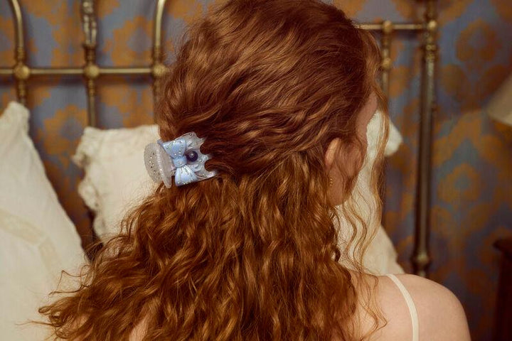 The Moonstone Ruku Hair Claw Rainbow in light blue acrylic is seen in curly hair, shown from behind in a bedroom environment. Model 3971