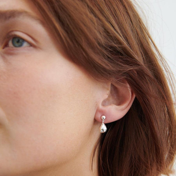 Pernille Corydon Driplet gold-plated earrings appear on the woman's ear in a glossy, drop-shaped style. Model E-306-GP-2