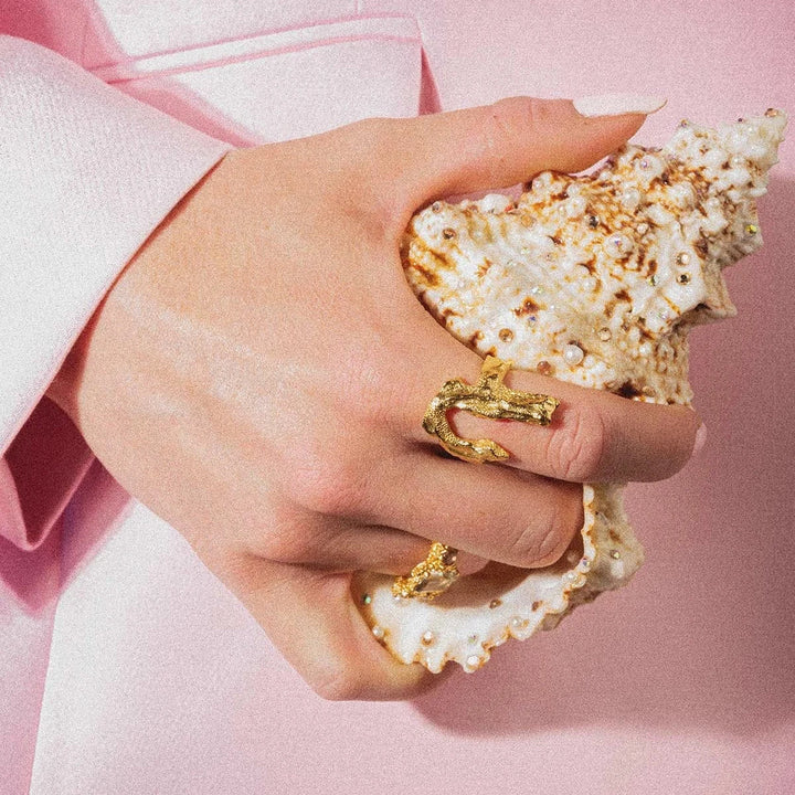 Vincent Bone Gold Plated Ring shown on hand, detail of gold colored ring against a conch shell. Model vj229-urg-001