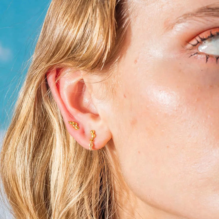 Vincent Pendulum Earrings Gold-plated shown on ear, small balls in gold look under clear sky. Model VJ177-UEG-2