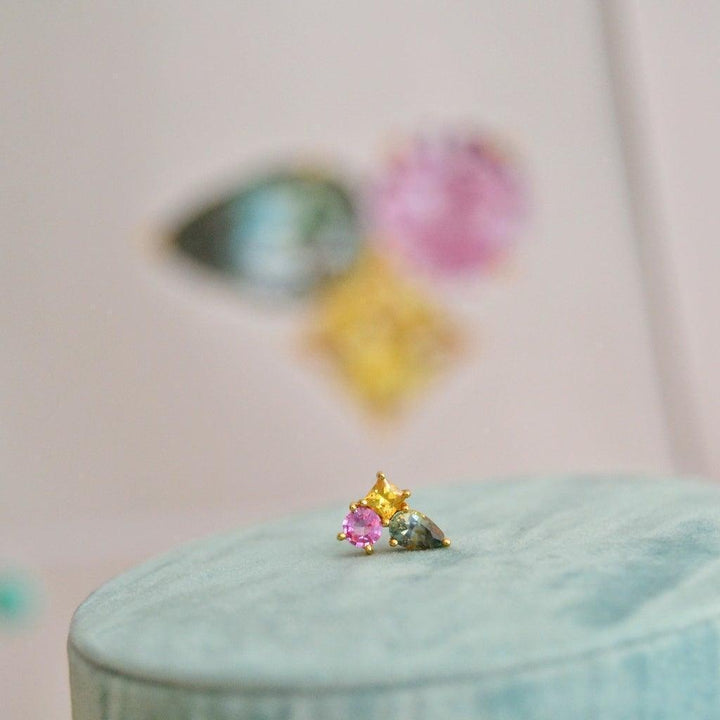 anpé atelier unika varnaya stud with sapphire 23 shows a colorful stud in gold with pink, yellow and blue stones on light green cushion. Model 23.